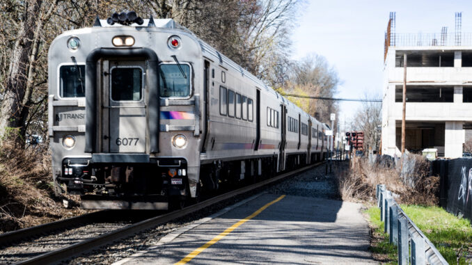 A commuter train wails its approach to Emerson Station downtown on April 13, 2024, arresting road traffic. The borough's mixed-use-redevelopment project, at right, on Kinderkamack between Lincoln and Linwood, languishes under construction with its principals tied up on cash flow and legal issues, some with the borough. John Snyder photo.