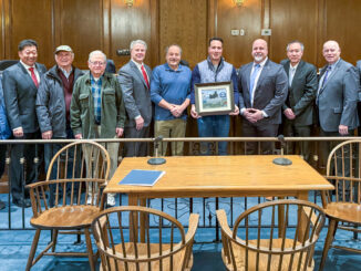 U.S. Marine Corps veteran Fred Puglisi (center, blue shirt), Old Tappan’s veterans organizer and chairman, stands with Peter Lugo, a recently retired U.S. Army colonel with the New Jersey State Veterans Chamber of Commerce, holding the certification plaque, and Mayor Thomas Gallagher (right). Also pictured are Old Tappan councilmembers and three honored Old Tappan veterans. The plaque includes a photo taken over Afghanistan by Col. Lugo of a Chinook helicopter carrying steel from the World Trade Center.