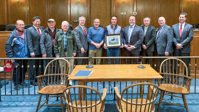 U.S. Marine Corps veteran Fred Puglisi (center, blue shirt), Old Tappan’s veterans organizer and chairman, stands with Peter Lugo, a recently retired U.S. Army colonel with the New Jersey State Veterans Chamber of Commerce, holding the certification plaque, and Mayor Thomas Gallagher (right). Also pictured are Old Tappan councilmembers and three honored Old Tappan veterans. The plaque includes a photo taken over Afghanistan by Col. Lugo of a Chinook helicopter carrying steel from the World Trade Center.
